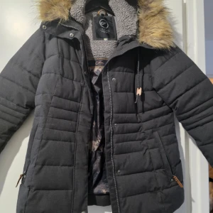 Grå dunjacka från Five Seasons knappt använd - Säljer en svart dunjacka från Five Seasons med fuskpäls på huvan och mjukt foder. Jackan har dragkedja och knappar framtill samt praktiska fickor med dragkedja. Perfekt för kalla dagar! 