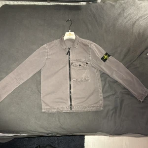 Grå overshirt från Stone Island - Säljer en snygg grå overshirt från Stone Island. Den har en dragkedja framtill och en praktisk bröstficka med knapp. Perfekt för höst och vår, med en stilren look som funkar både till vardags och fest. Märkeslogga på ärmen ger den extra stilpoäng. Passar perfekt för dig som gillar en avslappnad men ändå trendig stil. Liten skada på armen hör av er vid frågor