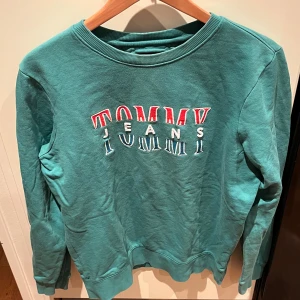 Grön sweatshirt från Tommy Hilfiger - Säljer en snygg grön sweatshirt från Tommy Hilfiger med broderad logga i rött och vitt på bröstet. Tröjan har en normal passform och är perfekt för både höst och vår. Den är i mycket bra skick och superbekväm att ha på sig. Hör av dig vid intresse 