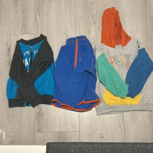 Färgglada barntröjor i olika stilar - Säljer tre coola barntröjor! En grå hoodie med texten 'PLAY TIME' i färgglada bokstäver, med orange huva och ficka. En blå fleecejacka med orange dragkedja från Quechua, perfekt för utomhuslek. Och en blå och grå zip-tröja med texten 'ATHLETIC TRAINING DEPARTMENT'. Alla i bra skick utan skador