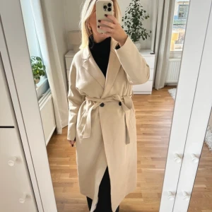 Beige trenchcoat med bälte - Intressebolag på en stilren beige trenchcoat i mycket bra skick. Den har ett bälte i midjan och knappar framtill. Perfekt för höst och vår! Passar till både vardag och fest! Skulle säga att den passar Xs-M