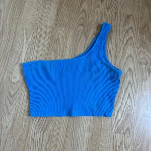 Blå ribbad oneshoulder topp - Säljer en snygg blå ribbad oneshoulder topp. Perfekt för sommaren med sin coola design och bekväma passform. Toppen är i ett mjukt material som känns skönt mot huden. Passar både till vardags och festliga tillfällen. Ett måste i garderoben! 💙