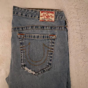 Blå jeans från True Religion - Säljer ett par snygga bootcut blå jeans från True Religion. De har en klassisk femficksdesign med slitna detaljer på bakfickan. Jeansen är i bra skick och har en bekväm passform. Perfekta för en avslappnad stil!