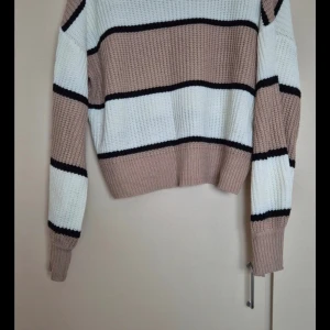Randig stickad tröja i beige och vit - Superfin stickad tröja med breda ränder i beige, vit och svart. Perfekt för kyliga dagar med sin mysiga och varma känsla. Tröjan har långa ärmar och en normal passform. Den ribbade strukturen ger en extra touch av stil. Passar perfekt till både jeans och kjol!