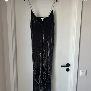 Svart glittrig långklänning - Säljer en elegant svart långklänning med glittriga paljetter. Perfekt för nyårsfesten eller en glamorös kväll. Klänningen har en smickrande passform och tunna axelband. Den är i mycket bra skick och redo att bäras av någon som vill glänsa!