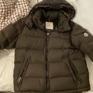 Säljer en snygg brun pufferjacka från Moncler i mycket bra skick. Jackan har en quiltad design med dragkedja och knappar, samt en mysig huva. Perfekt för kalla vinterdagar! Den är långärmad och har elastiska muddar för extra värme. Passar både till vardags och festliga tillfällen. Modellen är en chevalier jacket från 2014, och köptes från merch sweden! Smått slitage längst ut på ärmarna samt en del som saknas på zippern, inga problem att stänga och öppna dock!