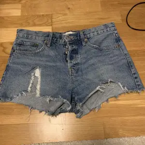 Säljer ett par coola blå jeansshorts med slitna detaljer och fransig kant. Perfekta för en avslappnad sommarlook. De har en klassisk femficksdesign och knappgylf. Passar perfekt för festivaler eller en dag på stranden!