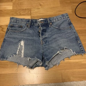 Blå jeansshorts med slitningar - Säljer ett par coola blå jeansshorts med slitna detaljer och fransig kant. Perfekta för en avslappnad sommarlook. De har en klassisk femficksdesign och knappgylf. Passar perfekt för festivaler eller en dag på stranden!