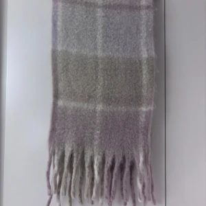 Rutig halsduk med fransar - Säljer en supermysig rutig halsduk i mjuka toner av beige och lila. Den är perfekt för kyliga dagar med sitt varma och sköna material. Halsduken har fransar i ändarna som ger en extra touch av stil. Perfekt för höst och vinter!