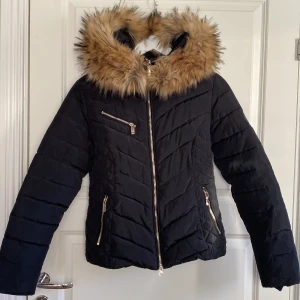 Svart dunjacka med pälsdetaljer från Roco Baroco - Säljer en snygg svart dunjacka från Roco Baroco i storlek 34, XS. Jackan har en fluffig pälsdetalj på huvan, pälsdetaljen är avtagbar och flera praktiska dragkedjefickor. Perfekt för kalla vinterdagar med sitt varma foder. Som ny.