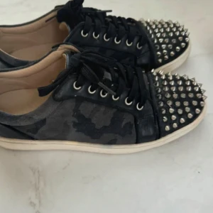 Svarta Christian Louboutins - Säljer ett par coola svarta sneakers med nitar på tån och en camouflagemönstrad design. Skorna har snörning och en röd sula som ger en extra touch. Perfekta för att ge din outfit en edgy stil!