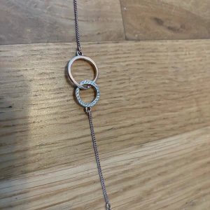 Elegant armband med dubbla ringar - Snyggt armband från Pilgrim med två sammanlänkade ringar. Den ena ringen är slät och den andra är dekorerad med gnistrande stenar. Armbandet har en tunn kedja som ger en stilren look. Perfekt för både vardag och fest!