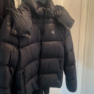 Svart dunjacka från Calvin Klein Jeans - Säljer en snygg svart dunjacka från Calvin Klein Jeans i storlek M. Jackan har en hög krage och dragkedja framtill, perfekt för kalla dagar. Den är i mycket bra skick och har en stilren design med logotyp på bröstet. Perfekt för både höst och vinter! 