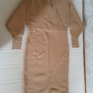 Beige klänning med pärldetaljer - Säljer en elegant beige klänning med pärldetaljer längs en omlott-design. Klänningen har långa ärmar och är gjord i ett mjukt material som känns skönt mot huden. Perfekt för höst och vår, och kan stylas både upp och ner beroende på tillfälle. Passar bra för festliga tillfällen eller en dag på stan.
