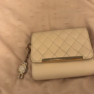 Handväska med robotberlock💘 - Säljer en superfin beige hand väska,men man kan lägga en kedja. Den har en söt liten robotberlock som hänger på sidan.Handväskan  är i mycket bra skick och har en stilren design med en metallisk detalj på framsidan.💘