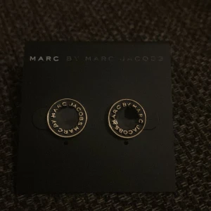 Svarta örhängen från Marc by Marc Jacobs - Snygga svarta örhängen från Marc by Marc Jacobs med guldtext runt kanten. Perfekta för att ge en stilren touch till vilken outfit som helst. De är i nytt skick och kommer i originalförpackning. Passar både till vardag och fest!