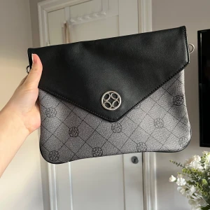 Svart och grå kuvertväska - Snygg kuvertväska i svart och grått med ett elegant mönster. Axelband tillkommer även. Vet inte original priset men den är köpt på Lundbergsväskor för ett par år sedan.