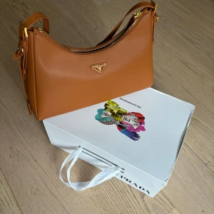  Prada Aimee  - Säljer en superfin brun axelväska från Prada modell Aimee. Väskan har en elegant design med guldfärgade detaljer och en praktisk dragkedja. Perfekt för både vardag och fest! Den är i mycket bra skick och kommer med originalförpackning.
