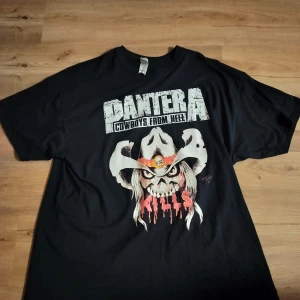 Svart Pantera T-shirt - Säljer en svart T-shirt med Pantera-tryck 'Cowboys from Hell'. Framsidan har en cool design med en dödskalle i cowboyhatt och blodiga detaljer. Baksidan listar turnéstäder med en stor dödskalle. Perfekt för rockfans! 🖤