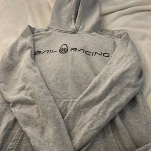 Grå hoodie från Sail Racing - Säljer en snygg grå hoodie från Sail Racing i storlek 150. Den har en klassisk design med en stor ficka fram och en bekväm huva. Perfekt för kyliga dagar eller som ett extra lager. Den är i bra skick och redo för nya äventyr!