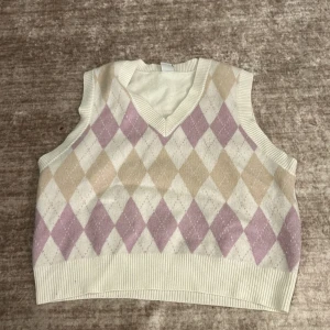 Rutig stickad väst från Lindex - Säljer en superfin stickad väst från Lindex i storlek 158/164. Den har ett klassiskt rutigt mönster i beige och lila med en v-ringad hals. Perfekt för lager-på-lager under hösten eller våren. Västen är i mycket bra skick. Säljer för att jag inte har haft på mig den sen 2021
