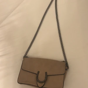 Beige axelväska med kedja - Säljer en snygg beige axelväska med en cool kedjerem. Väskan har en stilren design med ett dekorativt spänne på framsidan. Perfekt för både vardag och fest! 👜men är lite smutsig på baksidan 🤪💞