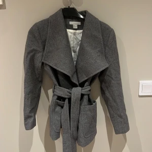 Grå kappa från H&M - Säljer en stilren grå kappa från H&M i storlek XS. Den har en elegant design med knytbälte i midjan och långa ärmar. Perfekt för höst och vår, och ger en chic look till vilken outfit som helst. Kappan är i mycket bra skick och redo för nya äventyr!
