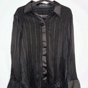 Svart glittrig skjorta från MNG - Snygg svart skjorta från MNG med glittriga ränder. Den har långa ärmar och en klassisk krage. Perfekt för festliga tillfällen! Skjortan är i bra skick och har en elegant look med knappar framtill.