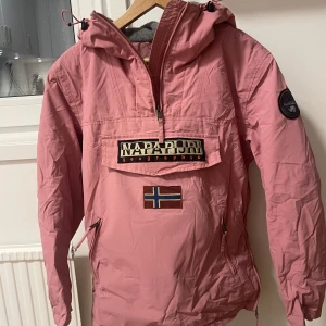 Rosa anorak från Napapijri - Säljer en snygg rosa anorak från Napapijri med en stor logga och norsk flagga på framsidan. Jackan har en praktisk dragkedja och en stor ficka framtill. Perfekt för höst och vår med sin långa ärmar och huva. Den är i bra skick och redo för nya äventyr! Liten i storleken så passar nog dam-small eller man-xs