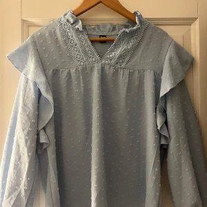 Ljusblå blus med spetsdetaljer - Säljer en superfin ljusblå blus från Sensity. Den har vackra spetsdetaljer vid halsen och volanger längs ärmarna. Blusen är långärmad och har ett diskret prickigt mönster. Perfekt för både vardag och fest!