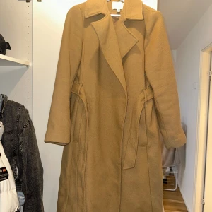 Beige kappa från H&M - Säljer en snygg beige kappa från H&M i storlek 36. Den har en klassisk design med ett bälte i midjan för en elegant passform. Perfekt för höst och vår även under vintern! 