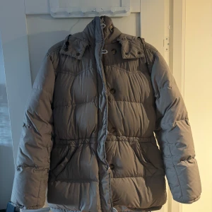 Grå pufferjacka med huva - Säljer en mysig och varm grå/beige pufferjacka perfekt för vintern. Jackan har en praktisk huva och stängs med både dragkedja och knappar för extra skydd mot kylan. Perfekt för kalla dagar! 
