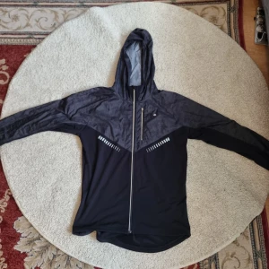 Svart vindjacka från Ronhill - Säljer en svart vindjacka från Ronhill i storlek M. Jackan har en huva och dragkedja framtill. Den är perfekt för löpning eller andra utomhusaktiviteter. Materialet är lätt och andas bra, vilket gör den idealisk för vår och höst. Reflexdetaljer på ryggen för extra synlighet i mörker.
