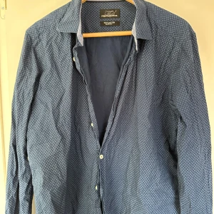 Blå slim fit skjorta från Finest Shirtmaker - Säljer en snygg blå skjorta från Finest Shirtmaker. Den är i 100% bomull och har ett diskret mönster som ger en stilren look. Skjortan är slim fit och har långa ärmar, perfekt för både vardag och fest. Knapparna framtill ger en klassisk touch. Passar perfekt till både jeans och kostymbyxor!