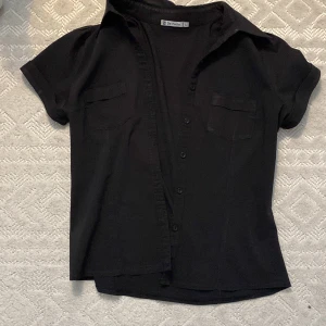 Svart kortärmad skjorta från De Facto - Säljer en stilren vintage svart kortärmad skjorta från De Facto, som jag fick av min mamma. Den är dock inte riktigt i min stil. Det står att storleken är L, men den ligger bra på mig som har storlek S. Perfekt att lägga över för den där sista touch.