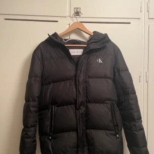 Svart pufferjacka från Calvin Klein storlek S - Snygg svart pufferjacka från Calvin Klein med huva och dragkedja. Jackan har en stilren design med CK-logga på bröstet och är perfekt för kyliga dagar. Använt skick men sparsamt använd.