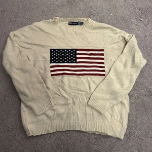 Beige stickad tröja från Faded Glory - Säljer en beige stickad tröja från Faded Glory med en stor amerikansk flagga på framsidan. Tröjan har långa ärmar och ribbstickade muddar vid ärmslut och nederkant. Perfekt för en avslappnad stil.