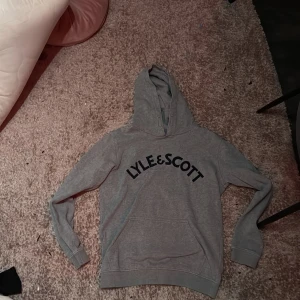 Grå hoodie från Lyle & Scott - Säljer en grå hoodie från Lyle & Scott med svart logga på framsidan. Perfekt för en avslappnad stil. Den har en klassisk huva och långa ärmar. Ord pris 649kr