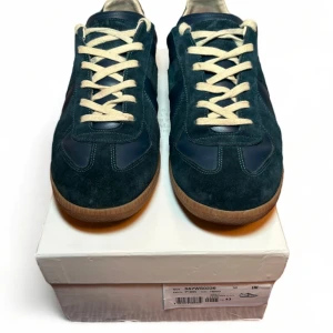 Maison Margiela GAT strl 43 - Snygga blåa sneakers från Maison Margiela med vit snörning och brun sula. Skorna är tillverkade i en kombination av mocka och skinn, vilket ger en stilren look. Perfekta för en avslappnad stil. Storlek 43. Gott skick, sparsamt använda av förra ägaren, men jag har inte använt dom alls eftersom de inte passade. Jag måste dock varna att om man är känslig mot parfym eller sånt så kan det vara bra att veta att de luktar mycket parfym från förra ägaren