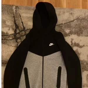 Svart och grå hoodie från Nike - Säljer en snygg svart och grå hoodie från Nike med dragkedja framtill. Perfekt för kyliga dagar med sin bekväma passform och stilrena design. Huvan ger extra värme och stil, den är köpt för 1200kr och är aldrig använd den är i top skick 