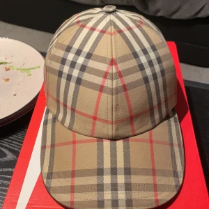 Burberry keps - Snygg rutig keps från Burberry i klassiskt beige, svart och rött mönster. Kepsen har justerbart spänne baktill med Burberry-logga. Perfekt för en stilren look. Helt nytt skick kvitto finns!!
