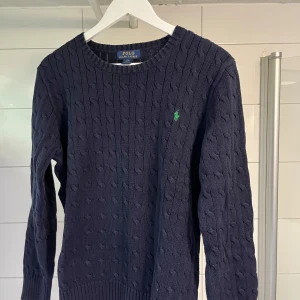 Mörkblå stickad tröja från Ralph Lauren - Säljer en snygg mörkblå stickad tröja från Ralph Lauren med kabelstickat mönster. Tröjan har en klassisk rund halsringning och det ikoniska gröna logotypbroderiet på bröstet. Tröjan är barnstorlek XL men passar de som har S eller mindre M. Mycket gott skick, knappt använd alls.
