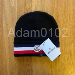 Svart mössa moncler mössa  - Snygg svart mössa från Moncler med röd och vit randdetalj. Den har Monclers logotyp broderad och är perfekt för kyliga dagar.