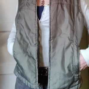 Grön dunväst med dragkedja - Säljer en stilren grön dunväst med dragkedja framtill. Västen har en hög krage och två praktiska fickor med dragkedjor. Perfekt för lager-på-lager under kyligare dagar.