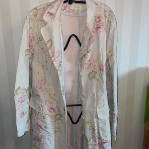 Blommig kappa från Marks&Spencer - Säljer en elegant blommig kappa /lång kavaj från Marks& Spencer. Den har ett vackert mönster med rosa och gröna blommor på en vit bakgrund. Kappan är långärmad och har knappar framtill. Perfekt för vår och sommar!