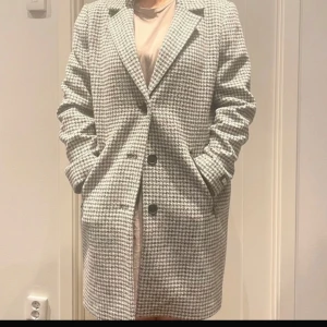 Rutig kappa från DKNY - Snygg rutig kappa från DKNY i svart och vit. Den har en klassisk design med knappar framtill och fickor med dragkedja. Perfekt för att hålla stilen under kyligare dagar.