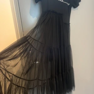 Svart klänning med text från Wiya - Trendig svart klänning från Wiya med texten 'smile' på bröstet. Klänningen har korta ärmar och en luftig, transparent kjol i flera lager. Perfekt för en stilren look.