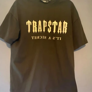 Svart t-shirt från Trapstar - Cool svart t-shirt från Trapstar med tryck fram och bak. Framsidan har texten 'TRAPSTAR IT'S A SECRET' i vitt, medan baksidan har 'PARIS' i en gradient av blått och orange. Perfekt för en avslappnad stil.