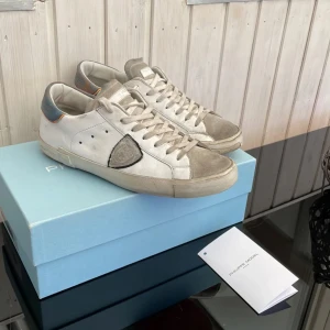 Vita sneakers från Philippe Model - Stilrena vita sneakers från Philippe Model med grå detaljer och snörning. Skorna har en klassisk design med en diskret logotyp på sidan och en bekväm sula. Perfekta för en avslappnad stil.