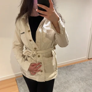 Beige jacka med knytbälte - Snygg beige jacka med knytbälte och knappar framtill. Perfekt för vår och höst. Jackan har långa ärmar och en stilren design som passar till många olika outfits. Den är ifrån Vero Moda, storlek S.